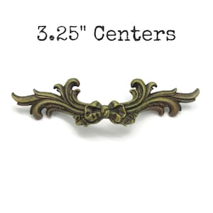 May include: An antique brass-toned metal drawer pull featuring an ornate, scrolling leaf design with a central bow detail. The text 3.25" Centers is printed above the hardware, indicating the distance between the mounting screw holes.