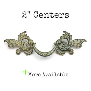 May include: Antique brass-toned drawer pull with ornate, scrolling leaf design. The pull has a curved handle and measures 2 inches between the centers of the mounting holes. The text "2" Centers" and "More Available" are also visible.