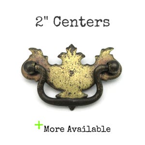 May include: Antique brass drawer pull with a decorative design. The pull has a curved handle and measures 2 inches between the centers of the mounting holes. The text "2" Centers" is above the pull.
