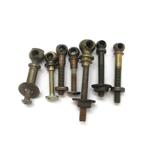 Peut inclure: Un ensemble de sept boulons à œil et fixations filetées en métal de longueurs et diamètres variés. Chaque pièce présente une tête en boucle circulaire et une tige filetée, avec un mélange de finitions en laiton, bronze et acier oxydé.
