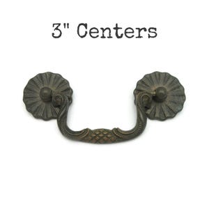 May include: Antique bronze drawer pull with a decorative floral design. The pull has a curved handle and measures 3 inches (7.6 cm) between the centers of the mounting holes. The text "3" Centers" is above the pull.