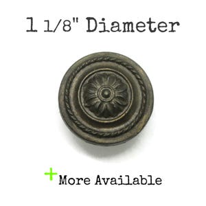 May include: A round, dark bronze-toned metal knob featuring a central floral rosette design surrounded by a rope-style border. The text 1 1/8" Diameter and + More Available is printed above and below the knob, indicating its size and availability.