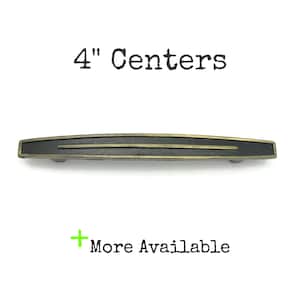 May include: A cabinet pull handle with a matte black center and antique brass-toned edges. The handle features a horizontal design with a thin brass line running through the middle. Text above reads 4" Centers and text below reads + More Available.