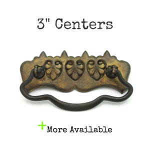 May include: Antique brass drawer pull with a decorative leaf design and a black handle. The text "3" Centers" is above the pull, and "More Available" is below. The pull measures 3 inches.