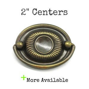 May include: Antique brass-toned drawer pull with a 2-inch center-to-center measurement. The oval-shaped pull features a central circular design surrounded by a ribbed pattern and two rounded end pieces. The text "2" Centers" and "More Available" are visible.