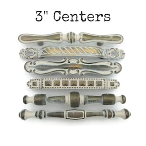 May include: A collection of six antique-style cabinet pulls with a distressed finish. The pulls are in various designs, including bar and handle styles, with colors of white, gold, and bronze. The text "3" Centers" is at the top of the image.