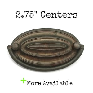 May include: An oval-shaped metal drawer pull with a dark, weathered finish and a central horizontal ridge. The hardware features a drop handle design with two mounting points spaced at 2.75" centers. The text 2.75" Centers and More Available is visible.