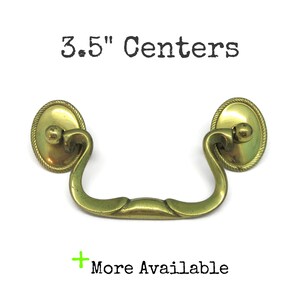 May include: A brass drawer pull with a curved handle and two oval mounting plates featuring a rope-style border. The text 3.5" Centers is displayed above the hardware, and the text More Available is positioned below with a green plus sign.