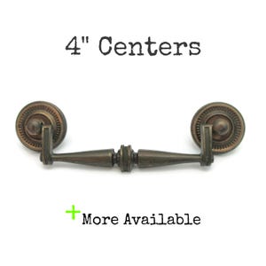 May include: Antique bronze cabinet pull with a 4-inch center-to-center measurement. The pull features a decorative circular backplate and a detailed handle. The text "4" Centers" is above the pull, and "More Available" is below.