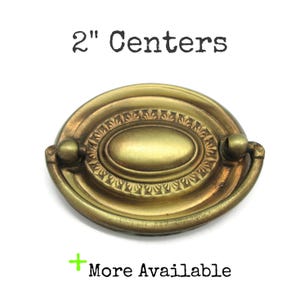May include: Oval-shaped brass drawer pull with a decorative center and two handles. The text "2" Centers" is above the pull, and "More Available" is below. The pull measures 2 inches across.