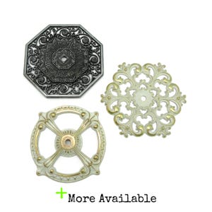 May include: Three decorative metal hardware pieces: one dark gray octagonal plate with intricate filigree, one cream and gold circular piece with a cross design, and one cream and gold floral-shaped piece. Text at the bottom reads: More Available.