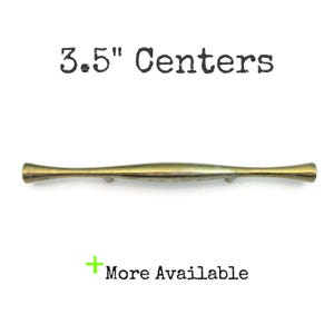 May include: A brass-toned cabinet pull with a curved, elongated design. The text "3.5" Centers" is above the pull, and "More Available" is below. The pull measures 3.5 inches in length.