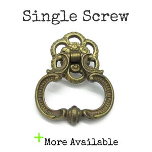May include: A brass-toned metal drawer pull with an ornate, openwork backplate and a swinging drop handle. The text "Single Screw" appears above the hardware, and the text "+ More Available" is positioned below the object.
