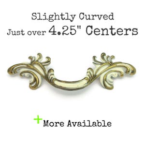 May include: A slightly curved, white and gold-toned cabinet pull with ornate floral detailing. The text reads "Slightly Curved Just over 4.25" Centers" and "More Available". This decorative hardware is ideal for furniture restoration projects.