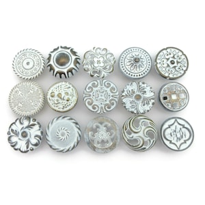 May include: A collection of fifteen decorative cabinet knobs in various designs. The knobs are primarily white with distressed, antique-style finishes in shades of gold and gray. Each knob features unique embossed patterns and textures.