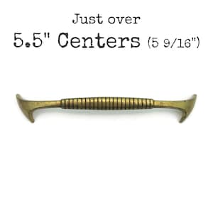May include: A brass-toned metal drawer pull with a ribbed center section and curved, pointed ends. The text above the hardware reads: Just over 5.5" Centers (5 9/16"). The handle features a weathered finish and a horizontal orientation.