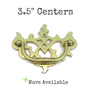 May include: A gold-toned brass drawer pull with a decorative openwork backplate and a curved drop handle. The text 3.5" Centers is printed above the hardware, and the text + More Available is printed below the hardware on a white background.