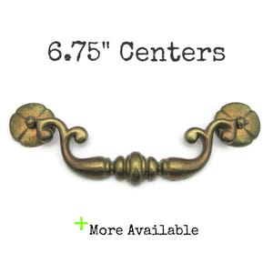May include: Antique brass drawer pull with a floral design on the ends. The pull measures 6.75 inches between the centers of the mounting holes. The text "6.75" Centers" is above the pull, and "More Available" is below.