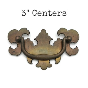 May include: A decorative antique brass drawer pull with a curved handle and ornate floral design. The text "3" Centers" is above the pull. The pull measures 3 inches between the mounting holes.