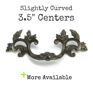 May include: A dark bronze-toned metal drawer pull with an ornate, scrolling leaf design. The text above reads Slightly Curved 3.5" Centers and the text below reads + More Available. The hardware features symmetrical, curved arms and a central detail.