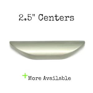 Peut inclure: Une poignée de meuble en métal argenté brossé avec une forme incurvée et allongée. Le texte 6,35 cm Centers est imprimé au-dessus de la quincaillerie, et un signe plus vert avec le texte More Available est imprimé en dessous sur fond blanc.