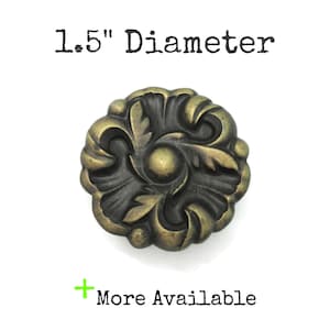 May include: A round metal cabinet knob with an antique brass finish and a raised floral leaf pattern. The knob has a 1.5 inch diameter. The text 1.5 inch Diameter and + More Available is printed in black above and below the knob.