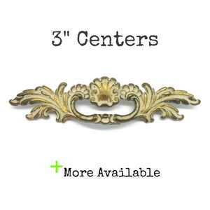 May include: A decorative metal drawer pull with an ornate, scrolling leaf and shell design, featuring a distressed cream and gold finish. The text 3" Centers is displayed above the pull, and the text + More Available is displayed below.