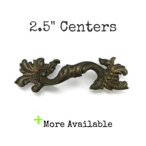 May include: An antique brass-toned metal drawer pull featuring ornate, scrolling leaf-like details on both ends. The text 2.5" Centers is printed above the handle, and a green plus sign with the text More Available is printed below the handle.