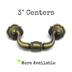 May include: A brass-toned metal drawer pull with a curved handle and two decorative mounting plates. The text 3" Centers is printed above the handle, and the text + More Available is printed below the handle in black lettering.