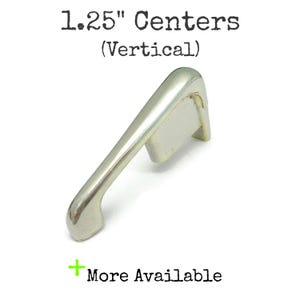 May include: A silver-toned metal cabinet pull with a smooth, curved handle design. The text above the object reads 1.25" Centers (Vertical) and the text below reads + More Available. The hardware features a solid construction with two mounting points.