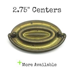 May include: An oval-shaped brass drawer pull with a weathered patina, featuring a central recessed oval detail and a swinging bail handle. The text 2.75" Centers and + More Available is printed in black above and below the hardware.
