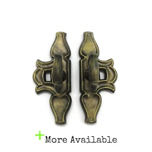 May include: Two antique brass-coloured metal fasteners with a vertical, ornate design. Each piece features a central rotating latch mechanism and a decorative, symmetrical frame with curved edges. The text "More Available" is printed below.