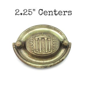 May include: Antique brass-toned drawer pull with a decorative oval center featuring a classical temple design. The pull has a 2.25-inch center-to-center measurement and is set against a white background.