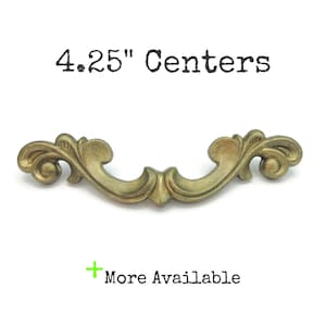 May include: A decorative brass-toned cabinet pull with an ornate, scrolling design. The pull measures 4.25 inches between the centers of the mounting holes. The text "4.25" Centers" is above the pull, and "More Available" is below.