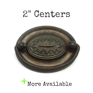 May include: An oval-shaped metal drawer pull with a dark, weathered bronze finish. The center features a raised geometric starburst pattern, and the outer edge has a braided texture. The text 2" Centers and + More Available is visible.
