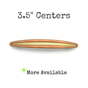 May include: A copper-colored cabinet pull with a gold-colored center. The text "3.5" Centers" is above the pull, and "More Available" is below it.