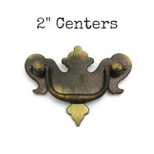 May include: Antique brass cabinet pull with a 2" centre to centre measurement. The pull has a decorative, ornate design with a curved handle.