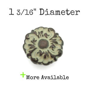 May include: A decorative cabinet knob with a floral design. The knob is 1 3/16" in diameter and features a distressed finish with cream and brown tones. The text "More Available" is also present.