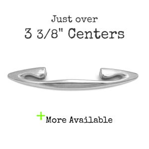 May include: A polished silver cabinet pull with a curved design. The text "Just over 8.5 cm Centers" is above the pull, and "More Available" is below. The hardware is designed for furniture and cabinetry.