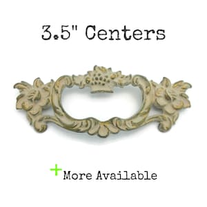 May include: A cream and gold antique-style drawer pull with floral and scrollwork details. The text "3.5" Centers" is above the handle, and "More Available" is below. The handle's center-to-center measurement is 8.89 cm.