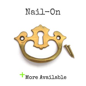 May include: A brass-toned nail-on drawer pull with a keyhole design and a curved handle. The text "Nail-On" is above the pull, and "More Available" is below. Two small nails are included.