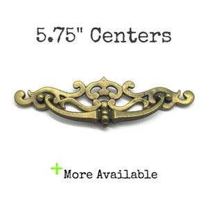 May include: An antique brass drawer pull with ornate scrollwork and a curved handle. The text "5.75" Centers" is above the pull, and "More Available" is below. The hardware is approximately 5.75 inches (14.6 cm) wide.