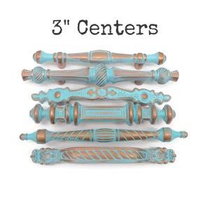 May include: Six drawer pulls with 3" centers, featuring a distressed turquoise and copper finish. Each handle displays a unique decorative shape with intricate molded details, showcasing a weathered metal appearance across all six hardware pieces.