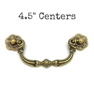 May include: Antique brass drawer pull with floral accents. The pull has a curved handle and measures 4.5" between the centers of the mounting holes. The text "4.5" Centers" is printed above the pull.