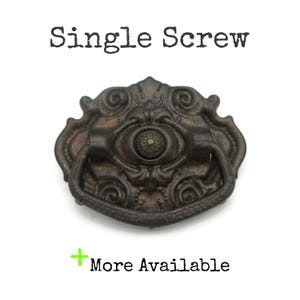 May include: Ornate, antique-style drawer pull with a dark bronze finish. The pull features a decorative oval center with a floral design and a curved handle. The text "Single Screw" is above the pull, and "More Available" is below.