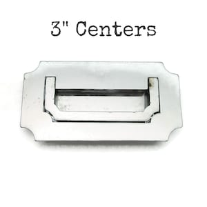 May include: A polished silver drawer pull with a rectangular handle and decorative edges. The text "3" Centers" is printed above the pull. The pull measures 3 inches between the centers of the mounting holes.
