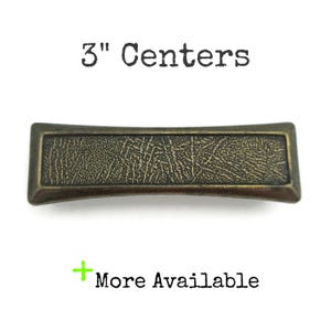May include: A rectangular cabinet pull with a textured surface and a bronze finish. The pull measures 3 inches (7.6 cm) between the centers of the mounting holes. The text "3" Centers" is above the pull, and "More Available" is below.
