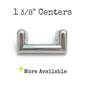 May include: A shiny, silver-toned cabinet pull with rounded edges. The text "1 3/8" Centers" is above the pull, and "More Available" is below. The hardware is designed for furniture and cabinetry.