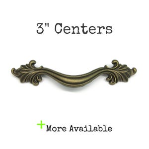 May include: An antique brass-toned metal drawer pull with a curved centre and ornate leaf-like ends. The text 3" Centers is printed above the handle, and the text + More Available is printed below the handle on a plain white background.