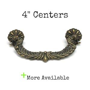 May include: An antique brass-colored metal drawer pull with ornate, curved, and leaf-like detailing. The handle features a central decorative element and two circular mounting ends. Text above reads 4" Centers and below reads + More Available.